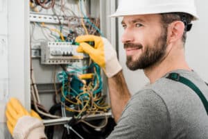 handsome cheerful electrician repairing electrical box and using screwdriver in corridor